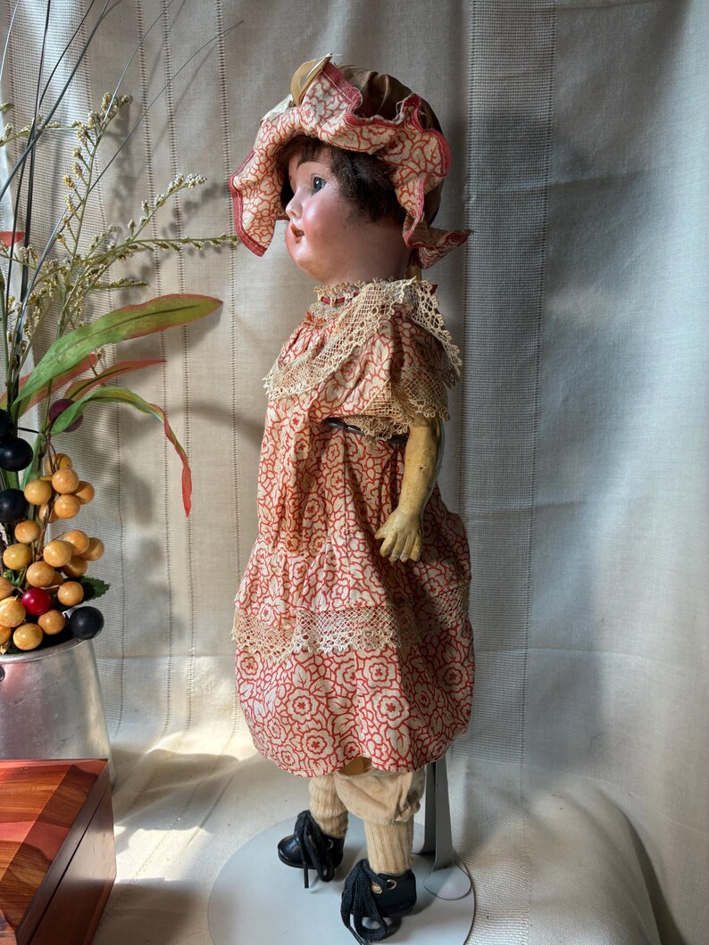 May include: A vintage doll with a painted face and dark hair, wearing a ruffled bonnet and a dress featuring a red and white floral pattern with lace trim. The doll has light-colored leggings and black shoes, standing on a metal display stand.
