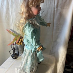 Op de afbeelding: Een vintage pop met blond haar, gekleed in een lichtblauwe jurk met een witte kanten rok. De pop heeft een porseleinen gezicht en staat in een zijaanzicht. De jurk heeft een satijnen afwerking en een bijpassende sjerp.
