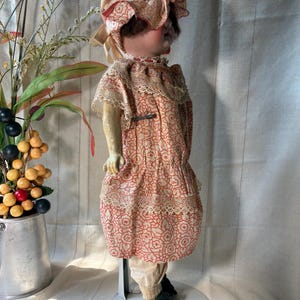 May include: A vintage doll wearing a peach-colored bonnet with a ruffled brim and a matching dress featuring a repeating red floral pattern. The dress is accented with cream-colored lace trim at the collar and waist. The doll wears light-colored trousers.