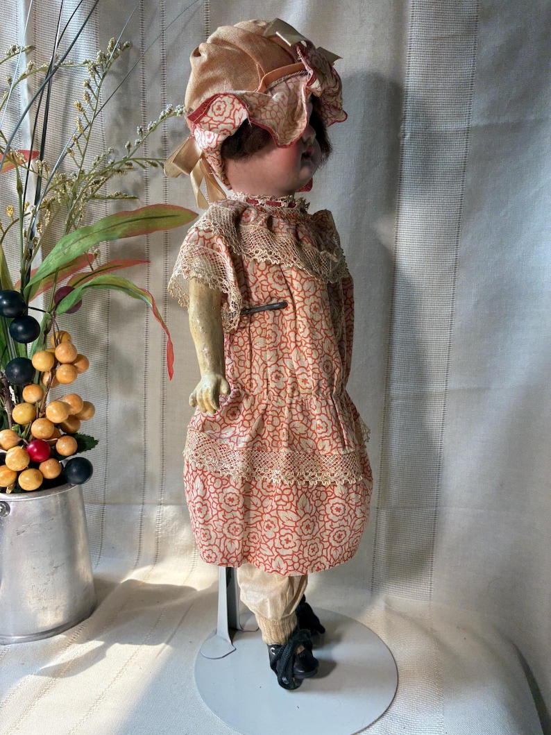 May include: A vintage doll wearing a peach-colored bonnet with a ruffled brim and a matching dress featuring a repeating red floral pattern. The dress is accented with cream-colored lace trim at the collar and waist. The doll wears light-colored trousers.
