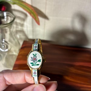 May include: Vintage gold-toned bracelet watch with a white face featuring a floral design. The watch has a white and gold band with floral accents. The watch face is oval-shaped with a gold bezel.
