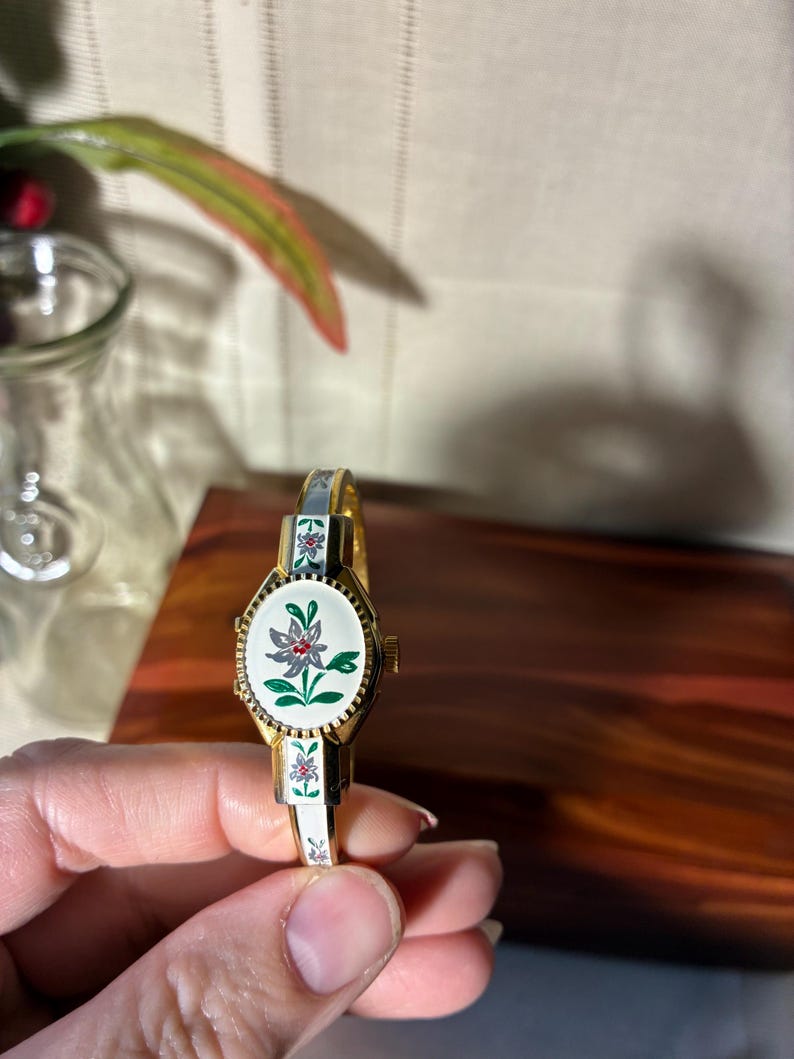 May include: Vintage gold-toned bracelet watch with a white face featuring a floral design. The watch has a white and gold band with floral accents. The watch face is oval-shaped with a gold bezel.