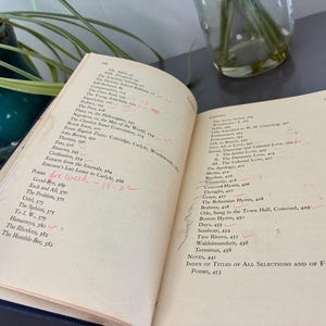 May include: An open book with aged, yellowed pages, featuring a list of poems and notes. Handwritten red markings and the text "Poems for week - 18-22" are visible. The book is held near a green ceramic pot with a plant.