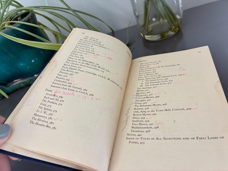 May include: An open book with aged, yellowed pages, featuring a list of poems and notes. Handwritten red markings and the text "Poems for week - 18-22" are visible. The book is held near a green ceramic pot with a plant.