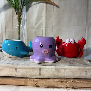 May include: Three ceramic animal-shaped planters: a blue whale, a purple octopus, and a red crab. The planters are sitting on top of a stack of old books. The whale has a small handle. The octopus has eight tentacles.