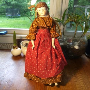 May include: A vintage doll with a porcelain face and a brown and red patterned dress. The doll's dress has a ruffled collar and sleeves, and a full skirt with white diamond-shaped patterns. The doll is standing on a wooden surface.