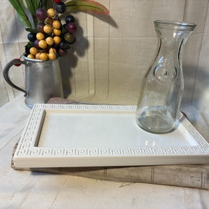 May include: A clear glass water carafe with a round emblem and a white rectangular serving tray with a Greek key design border. A metal pitcher with berries and a book are also present.