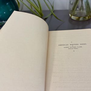 May include: An open book with off-white pages. The text "AMERICAN WRITERS SERIES" is visible, along with the author's name and "General Edition." The book is on a dark surface, with a glimpse of a green vase and plant in the background.