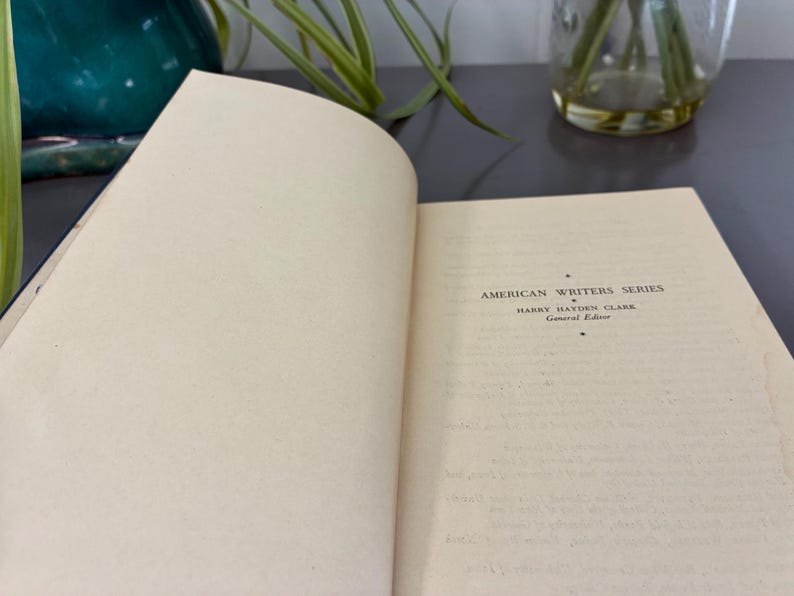 May include: An open book with off-white pages. The text "AMERICAN WRITERS SERIES" is visible, along with the author's name and "General Edition." The book is on a dark surface, with a glimpse of a green vase and plant in the background.