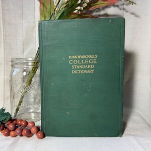 May include: A vintage green hardcover book titled "Funk & Wagnalls College Standard Dictionary" with gold lettering. The book is standing upright next to a clear glass jar and a sprig of red berries.