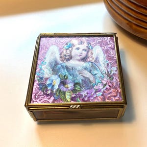 May include: A square brass trinket box with a hinged lid featuring a printed illustration of an angel with blonde hair and white wings, surrounded by blue and purple flowers against a textured pink background with a metallic gold frame.