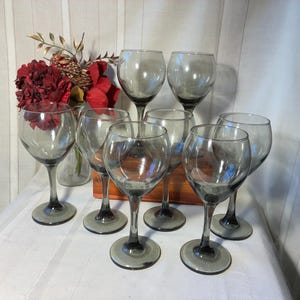 May include: A set of seven smoke-gray glass wine glasses with rounded bowls and stems. The glasses are arranged in a group, with some positioned behind others. A small glass vase containing red artificial flowers is visible in the background.