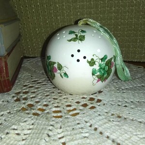 May include: A white ceramic ball-shaped tea infuser with a green ribbon. The infuser is decorated with green and pink floral designs and has small holes for steeping tea. A decorative item for tea lovers.