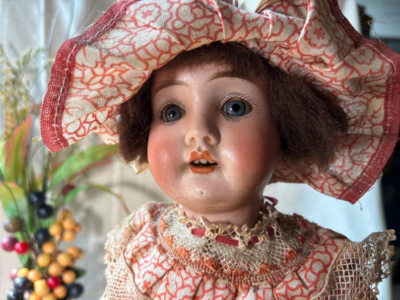 May include: A vintage doll with brown hair and blue eyes, wearing a wide-brimmed hat and a dress featuring a red and white floral pattern. The doll has painted facial features, including arched eyebrows, rosy cheeks, and orange-tinted lips.