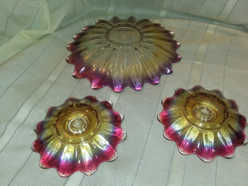 May include: Set of three iridescent glass dishes with a scalloped edge. The dishes are yellow, pink, and purple with a gold iridescent finish. The largest dish is a serving bowl, and the two smaller dishes are candle holders.