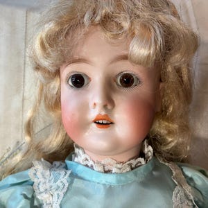 Op de afbeelding: Een vintage porseleinen pop met blond, krullend haar en bruine ogen. De pop heeft roze wangen en een kleine, open mond. Ze draagt een lichtblauwe jurk met een witte kanten kraag. Een klassiek voorbeeld van antiek speelgoed vakmanschap.