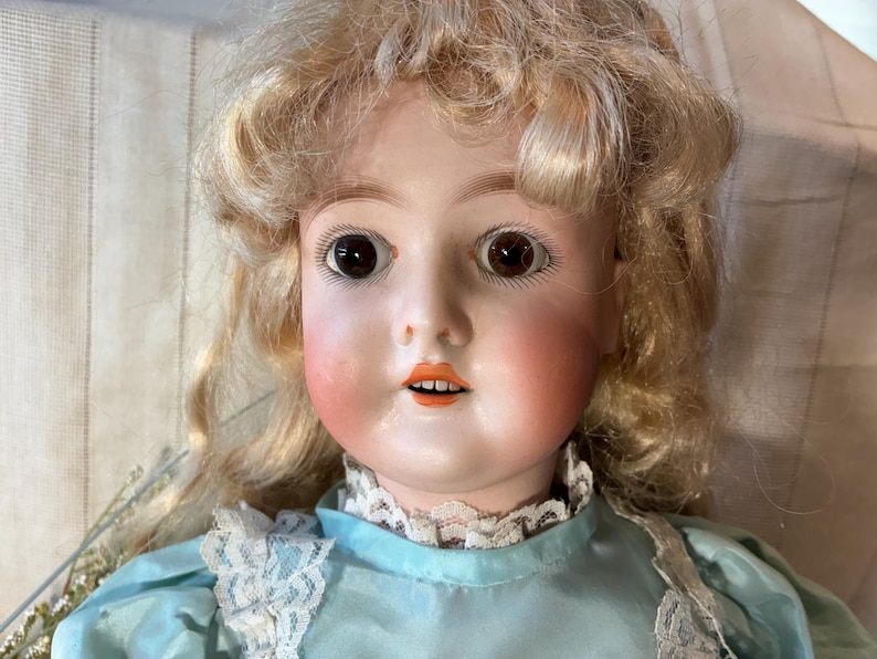 Op de afbeelding: Een vintage porseleinen pop met blond, krullend haar en bruine ogen. De pop heeft roze wangen en een kleine, open mond. Ze draagt een lichtblauwe jurk met een witte kanten kraag. Een klassiek voorbeeld van antiek speelgoed vakmanschap.