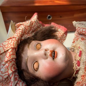 May include: A vintage doll with a pale face, rosy cheeks, and brown hair, wearing a ruffled bonnet and dress featuring a red and white floral pattern. The doll has orange-colored eyes, painted eyebrows, and an open mouth showing small teeth.