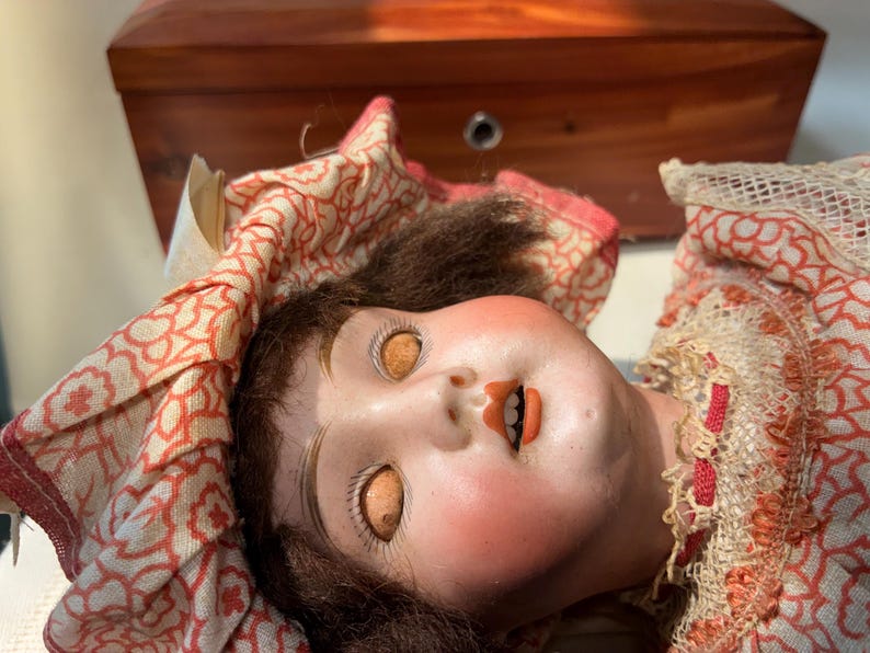 May include: A vintage doll with a pale face, rosy cheeks, and brown hair, wearing a ruffled bonnet and dress featuring a red and white floral pattern. The doll has orange-colored eyes, painted eyebrows, and an open mouth showing small teeth.