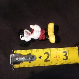 May include: A Mickey Mouse figurine, lying on its back, with a yellow measuring tape next to it. The figurine is black, white, red, and yellow. The measuring tape shows a measurement of approximately 2 inches.