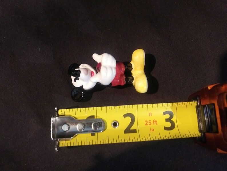 May include: A Mickey Mouse figurine, lying on its back, with a yellow measuring tape next to it. The figurine is black, white, red, and yellow. The measuring tape shows a measurement of approximately 2 inches.