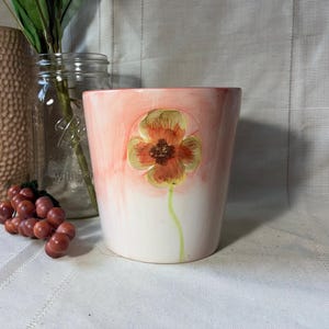 May include: A ceramic flower pot with a hand-painted orange and yellow flower design. The pot has a white base that fades into a peach-colored top. A green stem supports the flower.