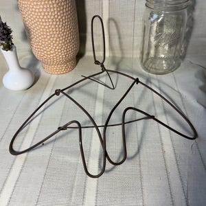 May include: A vintage-style metal wire hanger with a unique, abstract design. The hanger is dark brown and features a loop for hanging. A white vase with flowers, a textured peach-colored vase, and a glass jar are also in the image.