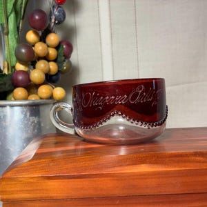 May include: A ruby red glass teacup with a clear glass handle and base. The cup is etched with the words "Niagara Falls" and has a beaded design around the rim. The cup sits on a wooden surface.
