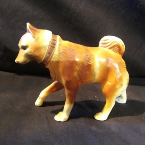 May include: A brown and white ceramic figurine of a dog with a brown leather collar. The dog is standing on all four legs and has its head tilted to the side.