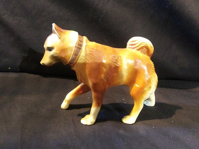 May include: A brown and white ceramic figurine of a dog with a brown leather collar. The dog is standing on all four legs and has its head tilted to the side.