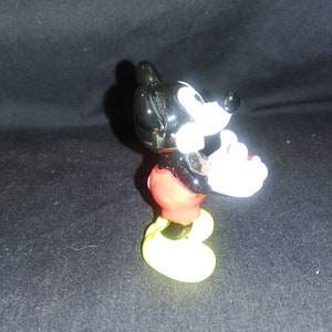 May include: A small figurine of Mickey Mouse, a classic cartoon character. The figure has a black head and ears, white gloves, red shorts, and yellow shoes. The figurine is posed with one hand raised.