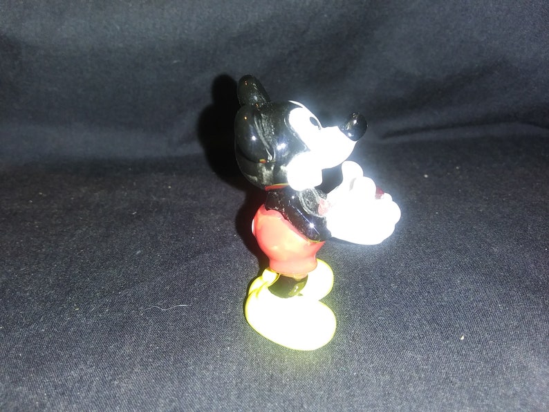 May include: A small figurine of Mickey Mouse, a classic cartoon character. The figure has a black head and ears, white gloves, red shorts, and yellow shoes. The figurine is posed with one hand raised.