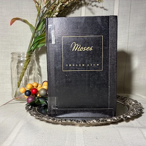 May include: A hardcover book titled "Moses" by Sholem Asch, displayed on a decorative silver-colored tray. The book has a black cover with gold lettering. A glass jar with flowers and decorative berries is in the background.
