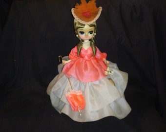 May include: A vintage doll with large eyes, wearing a pink dress with a white lace overlay and a matching orange hat. The doll is holding a gold-colored walking stick and a pink umbrella.