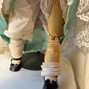 Op de afbeelding: Close-up van de benen van een antieke pop. De pop draagt zwarte schoenen met gouden accenten, witte sokken en een witte broek met kanten rand. De benen van de pop zijn van hout en vertonen tekenen van ouderdom.