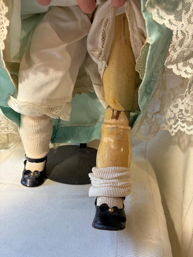 Op de afbeelding: Close-up van de benen van een antieke pop. De pop draagt zwarte schoenen met gouden accenten, witte sokken en een witte broek met kanten rand. De benen van de pop zijn van hout en vertonen tekenen van ouderdom.