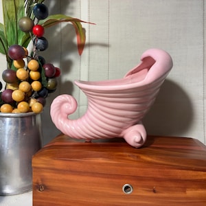 May include: A pink ceramic cornucopia vase with a curled end and ribbed texture. The vase sits on a wooden box. A metal pitcher with faux fruit and greenery is in the background.
