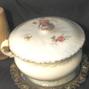 May include: A white ceramic tureen with a lid, featuring floral designs in pink and green. The tureen has a handle and sits on a decorative silver-colored tray. The lid has a brown knob. The rim of the tureen is trimmed with gold.