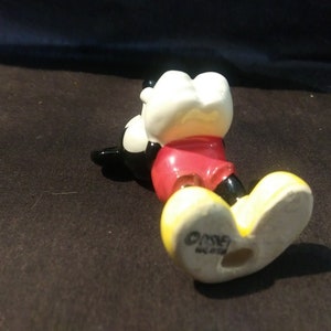 May include: A ceramic Mickey Mouse figurine, lying on its side. The figure has a white head, red shorts, black shoes, and yellow feet. The base of the foot is stamped with the Disney logo.