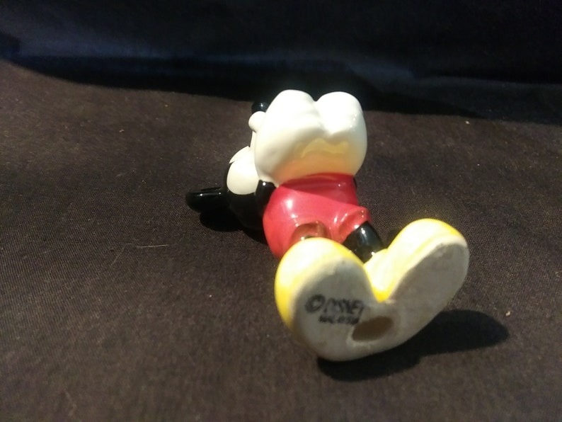 May include: A ceramic Mickey Mouse figurine, lying on its side. The figure has a white head, red shorts, black shoes, and yellow feet. The base of the foot is stamped with the Disney logo.