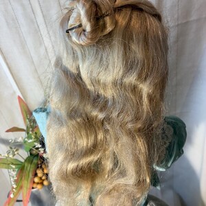 Op de afbeelding: Een vintage pop met lang, golvend blond haar in een updo. De pop draagt een lichtblauwe jurk met een groen lijfje. Het haar is vastgezet met donkere haarspelden. Het haar van de pop valt over de rug en cre&euml;ert een klassieke, elegante look.