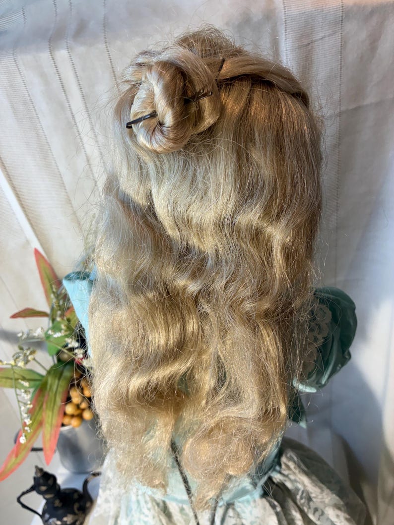 Op de afbeelding: Een vintage pop met lang, golvend blond haar in een updo. De pop draagt een lichtblauwe jurk met een groen lijfje. Het haar is vastgezet met donkere haarspelden. Het haar van de pop valt over de rug en cre&euml;ert een klassieke, elegante look.