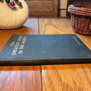 May include: A dark blue book titled "The Greatest Thing in the World" by Henry Drummond. The book is on a wooden surface. The title is in white text. A decorative white shell and a small woven basket are in the background.