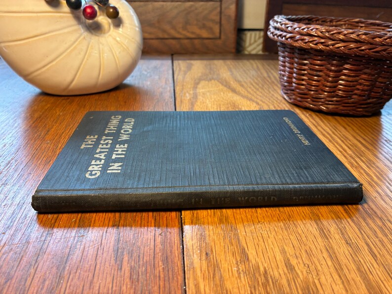May include: A dark blue book titled "The Greatest Thing in the World" by Henry Drummond. The book is on a wooden surface. The title is in white text. A decorative white shell and a small woven basket are in the background.