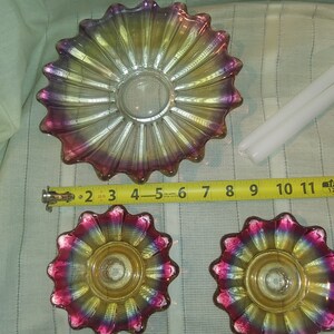 May include: Three iridescent glass candle holders with a scalloped edge and a yellow centre. The candle holders are on a white fabric surface with a measuring tape.