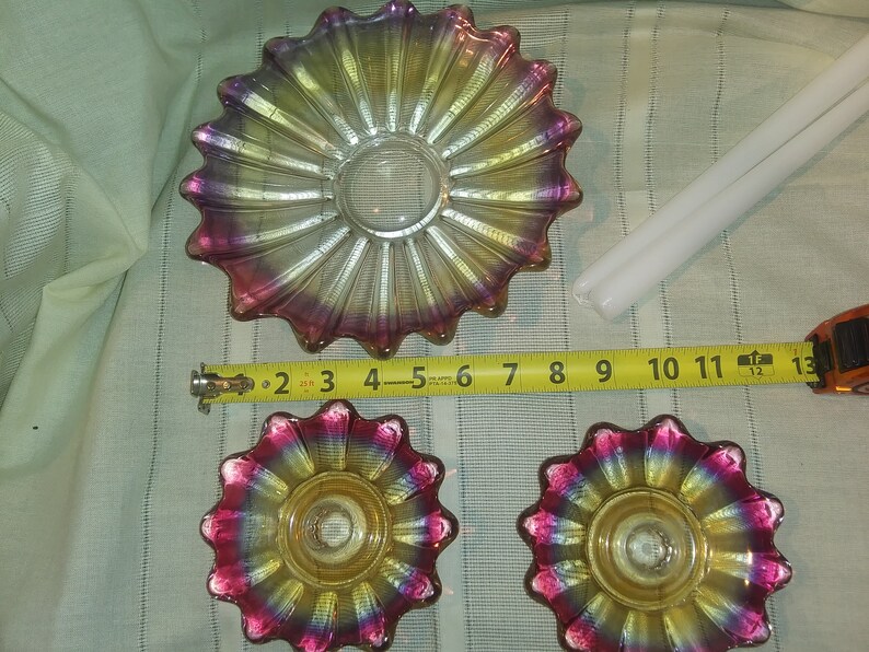 May include: Three iridescent glass candle holders with a scalloped edge and a yellow centre. The candle holders are on a white fabric surface with a measuring tape.