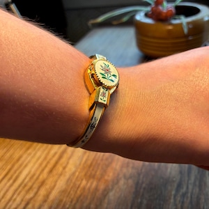 May include: Gold-toned hinged bracelet with a floral design. The bracelet features a central oval medallion with a painted floral motif in shades of red, green, and white. The band is white with gold accents and floral details. A vintage jewelry piece.