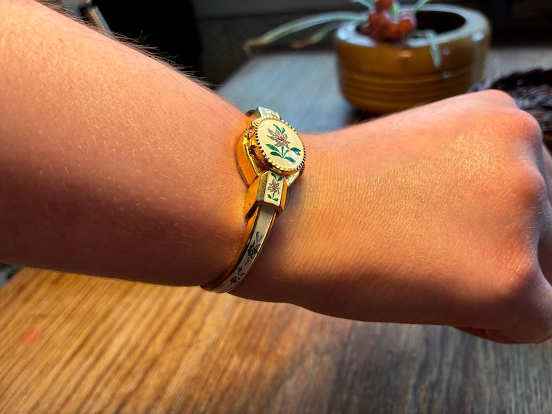 May include: Gold-toned hinged bracelet with a floral design. The bracelet features a central oval medallion with a painted floral motif in shades of red, green, and white. The band is white with gold accents and floral details. A vintage jewelry piece.