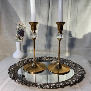 May include: Two gold-colored candlesticks with crystal accents and white candles sit on a silver-rimmed mirrored tray. A small white vase with dried flowers is to the left. The background is a white fabric.