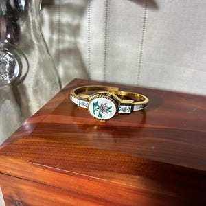 May include: Gold-toned hinged bracelet with a central oval design featuring a floral motif in pink and green on a white background. The bracelet has white enamel accents with floral details. A vintage jewelry piece.